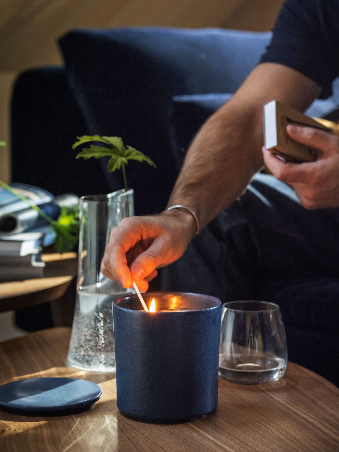 Iemand steekt een donkerblauwe FRUKTSKOG geurkaars aan met een lucifer. Op de achtergrond zie je een donkerblauwe bank.