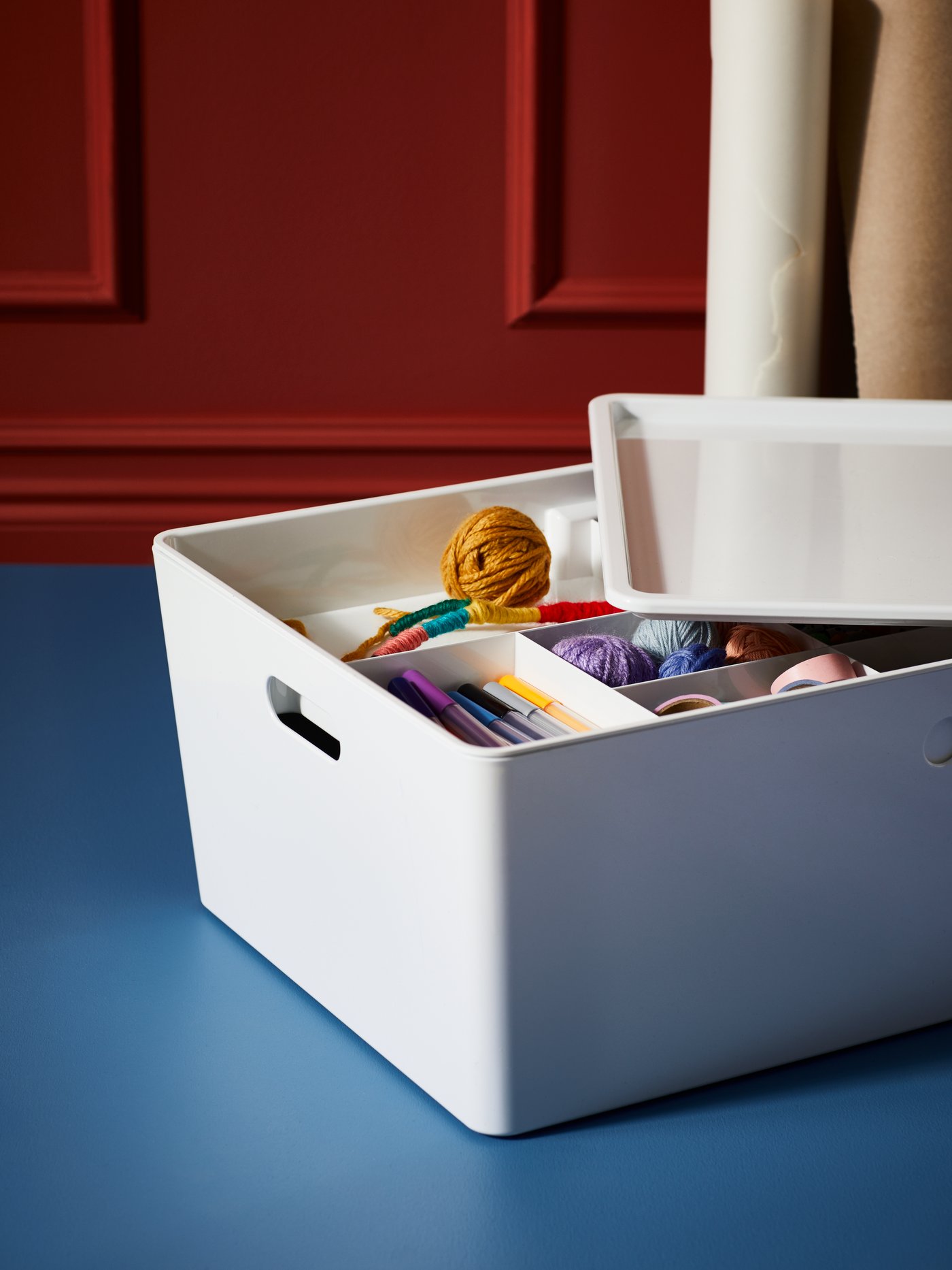 Una caja KUGGIS blanca sobre una mesa azul. Dentro de la caja hay una bandeja con compartimientos y algunos materiales de costura organizados.