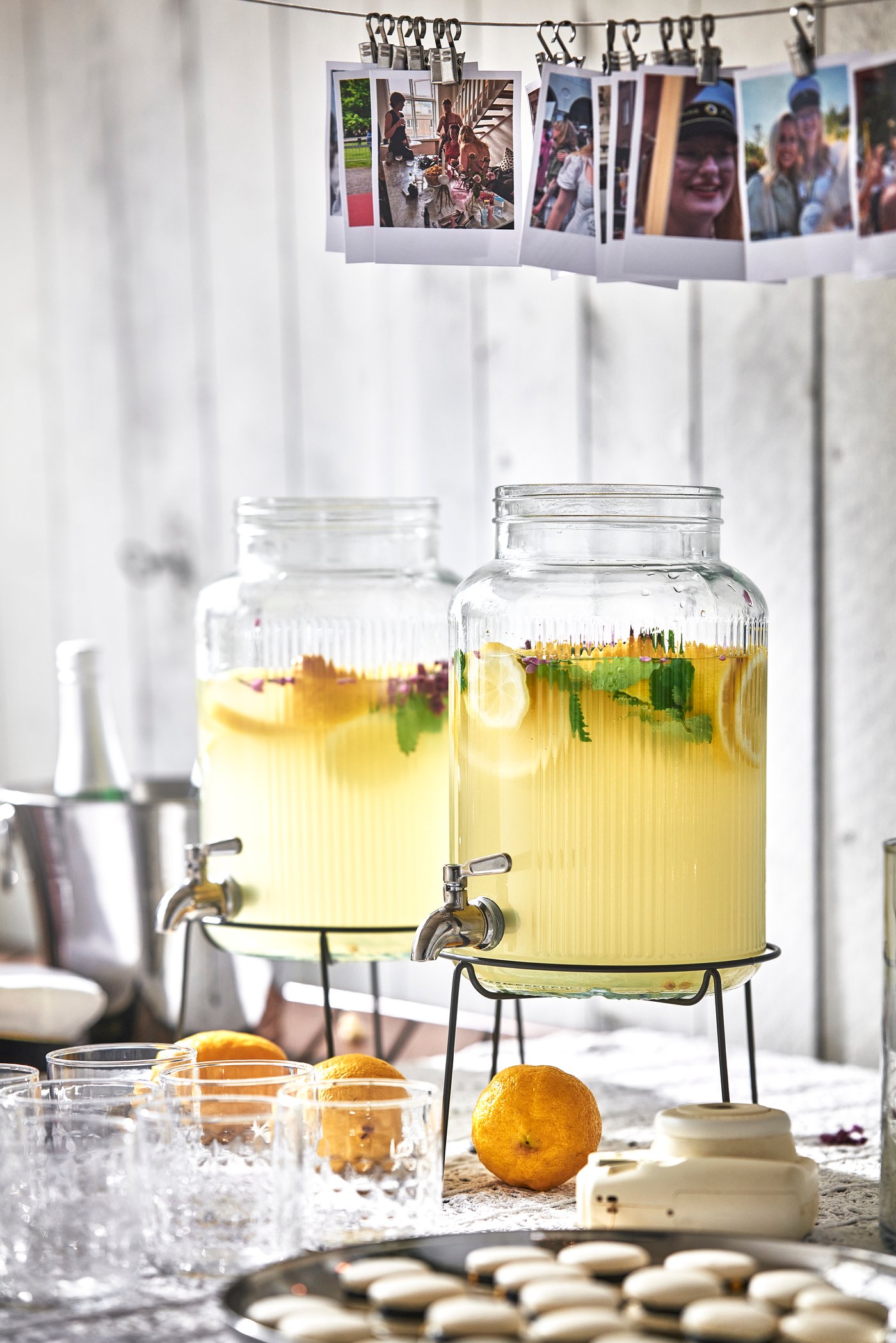 Två glasbehållare fyllda med ljusgul dryck med citrus och örter står på ett bord tillsammans med glas och apelsiner.