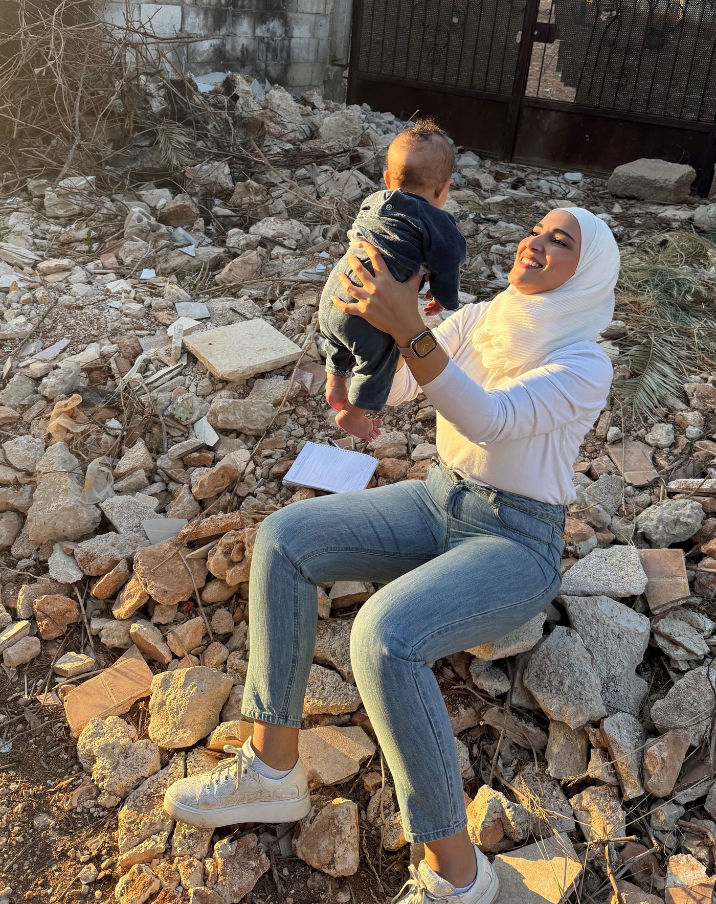 Lujain sitting outside on a pile of rocks holding a baby in her arms while smiling.