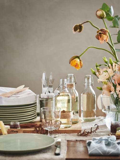 O secțiune a unei mese festive, cu un teanc de farfurii FÄRGKLAR verde mat, sticle KORKEN, pahare cu picior IKEA 365+ și flori.