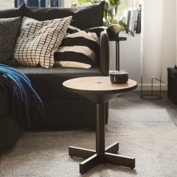 The round GRIMSARBO side table in black with a wooden top placed by a dark grey sofa with some black and white cushions.
