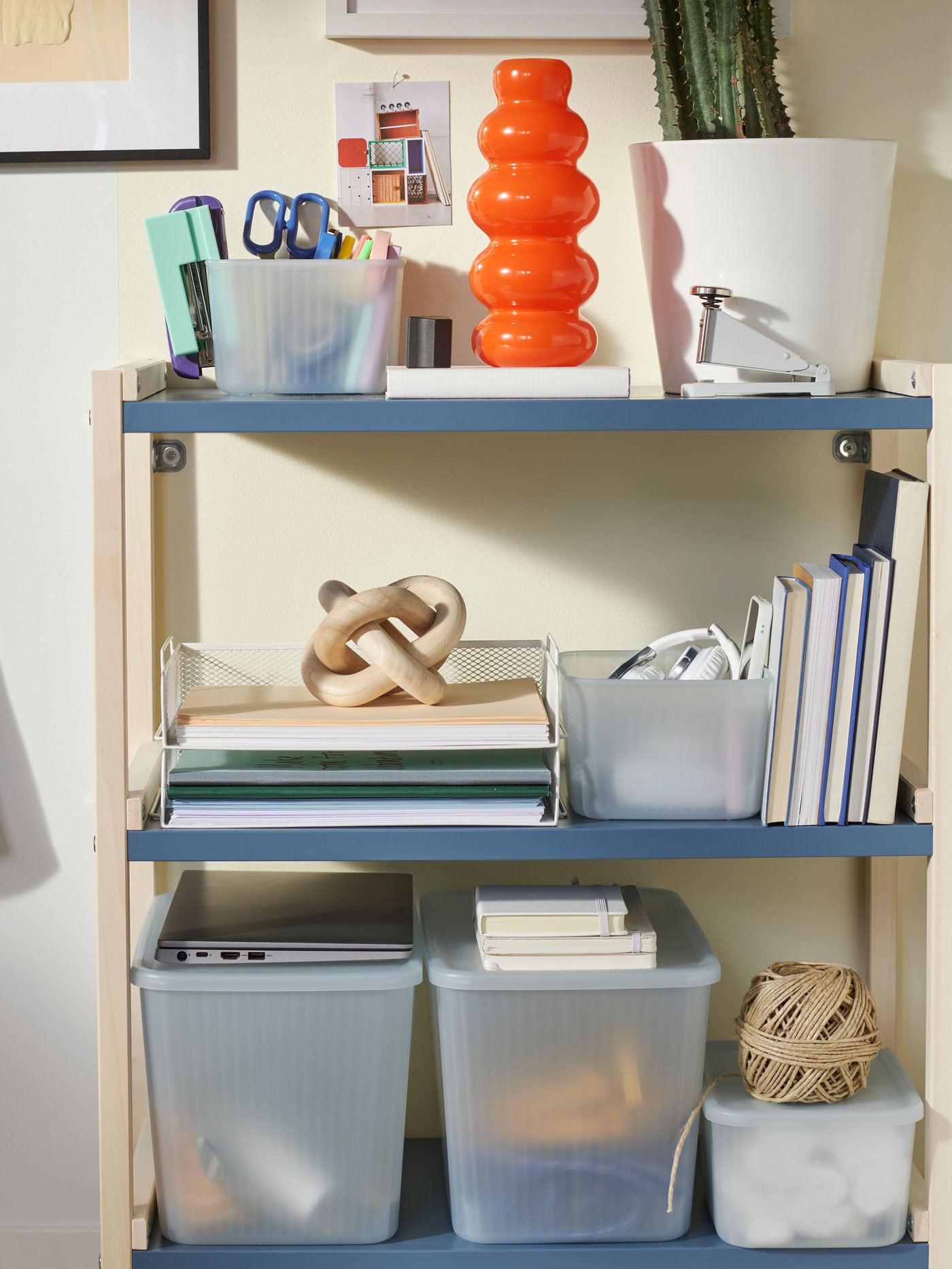 An open shelving unit with office supplies, storage boxes, a cactus, a white DRÖNJÖNS letter tray, and a curvy, orange vase.