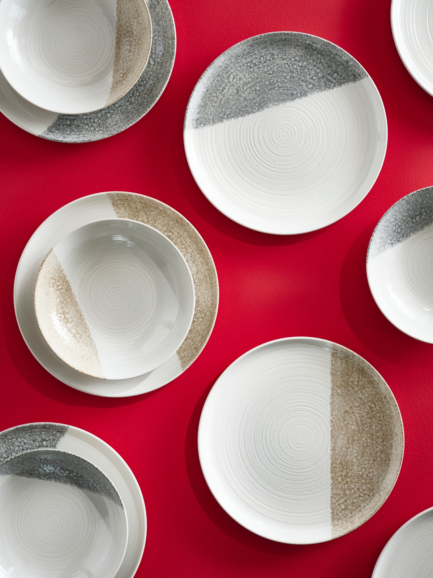 Vista dall'alto di diversi piatti piani e fondi KRUSTISTEL in grigio e beige chiaro, posizionati su una superficie rosso brillante.