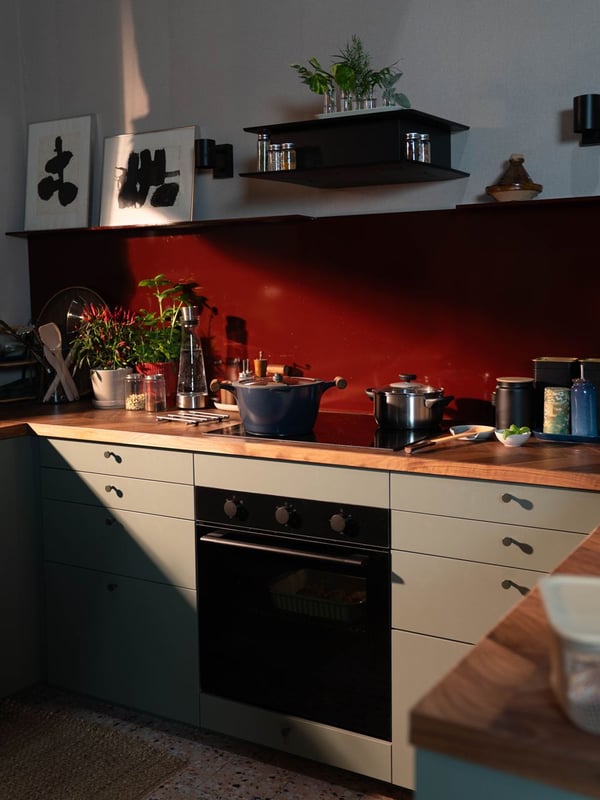 Una cucina con mobili color salvia e top in legno, illuminata da un raggio di sole che mette in risalto il paraschizzi rosso.