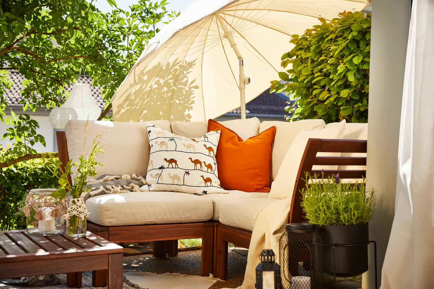 Gemütliche Outdoor-Sitzecke mit weissen Polstern, Zierkissen, Holzmöbeln und Sonnenschirm im grünen Garten.