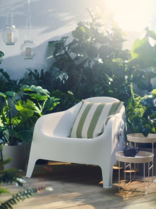 A white SKÄRPÖ outdoor armchair among lots of leafy green plants in pots and on plant stands, outside on a deck.