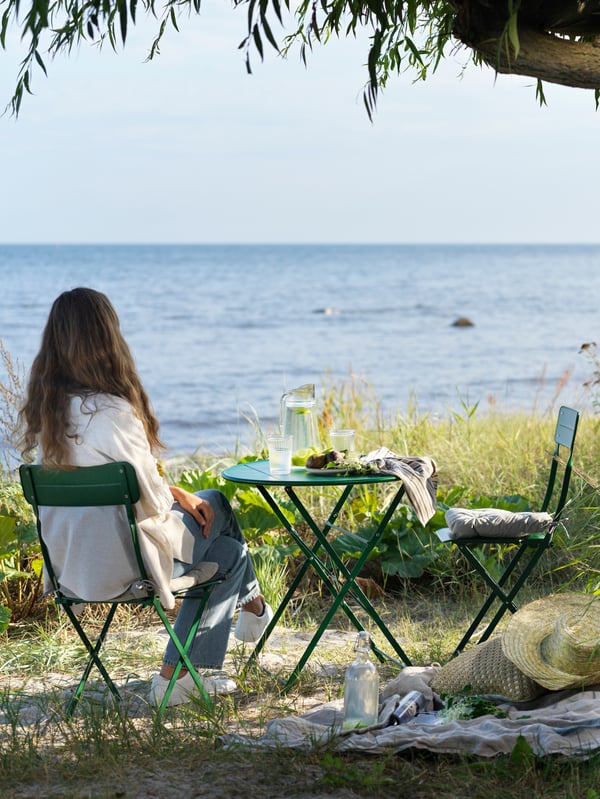 Una donna seduta di spalle che contempla il mare su una sedia all'aperto con un tavolino verde.