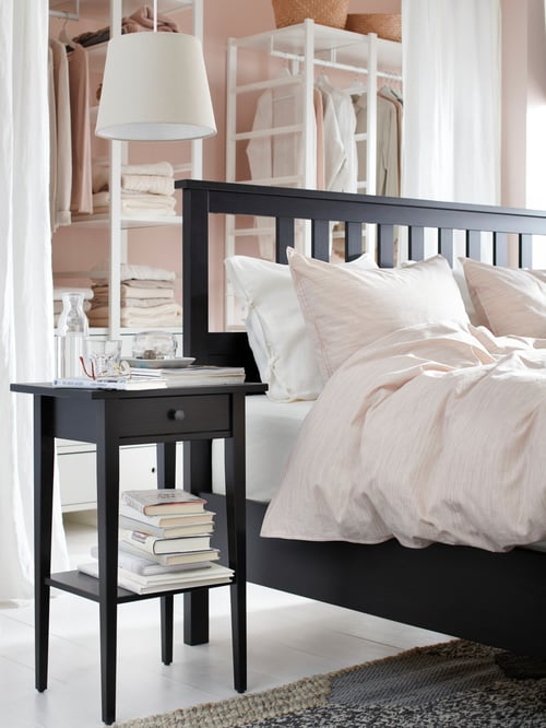 Cadre de lit HEMNES brun-noir installé devant trois éléments de rangement blancs, à côté d’une table de chevet noire avec des livres empilés.