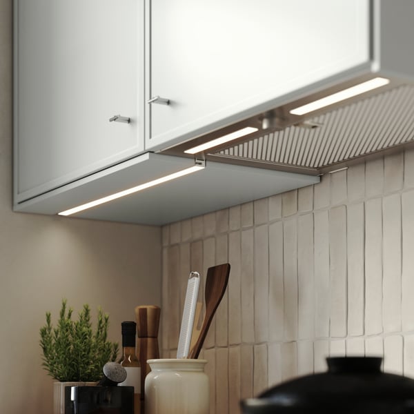 Ett kök med MITTLED LED-belysningslist för köksbänkskiva under ljusgrå skåp, som lyser mot den kaklade väggen och bänkskivan.