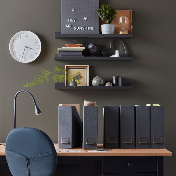 Auf einem Arbeitstisch stehen mehrere dunkle Ordner und eine schwarze Tischlampe. Darüber hängen 3 Regale mit Büchern und Bildern.