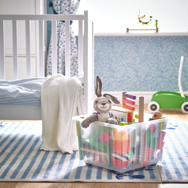 Una caja para juguetes VESSLA con ruedas incorporadas junto a una cuna SUNDVIK blanca. La caja es traslúcida y contiene un peluche de conejo y juguetes de madera MULA.