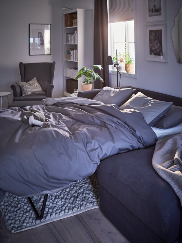 A dark grey GRÖNLID three seat sofa bed made up as a bed with grey bedding with the sun shining in under the half-open blind.