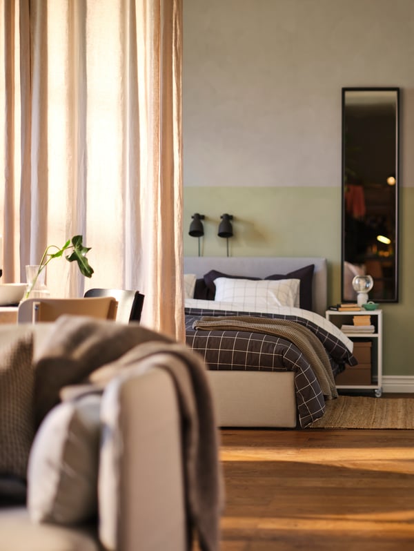A room-diving curtain separating a sleep area with a light grey GLADSTAD upholstered bed frame from a dining and living area.