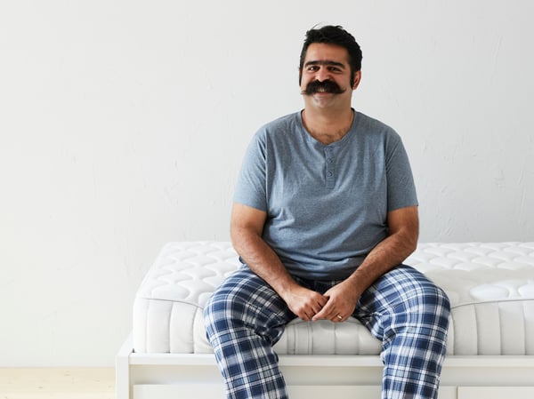A man with a moustache and wearing a greyish-blue t-shirt and blue plaid pyjama pants sits on the edge of a mattress.