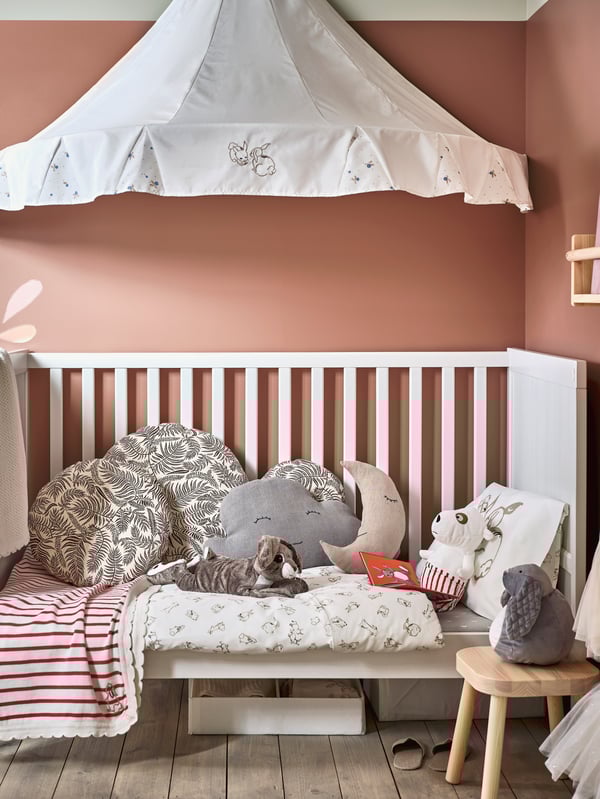 Ein Mädchenzimmer mit Wänden in Rosa und einem weißen Babybett mit vielen kuscheligen Textilien und Stofftieren und einem weißen Betthimmel darüber.