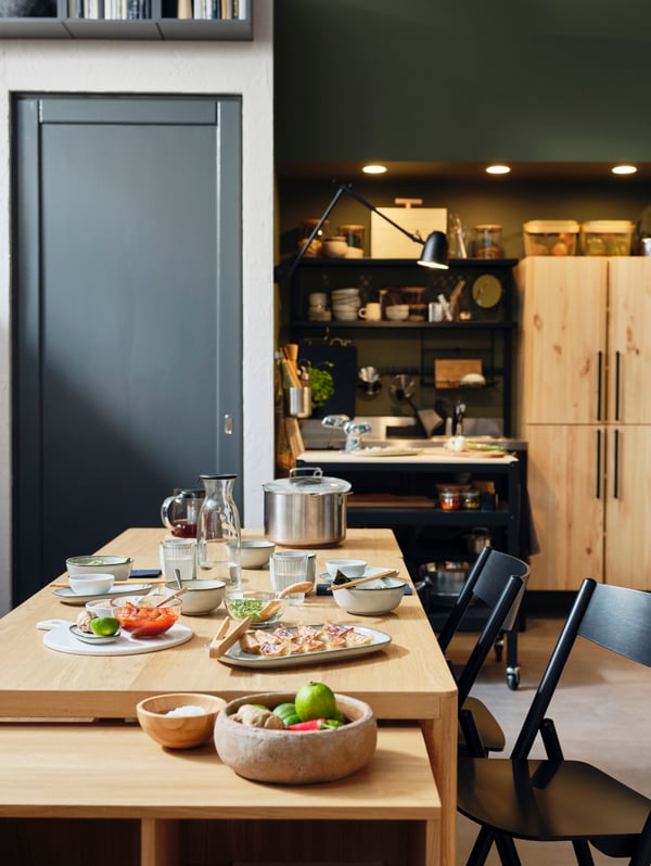 Una mesa, un módulo de almacenaje y unas sillas plegables RÅVAROR en medio de un apartamento. Encima de la mesa hay una vajilla.