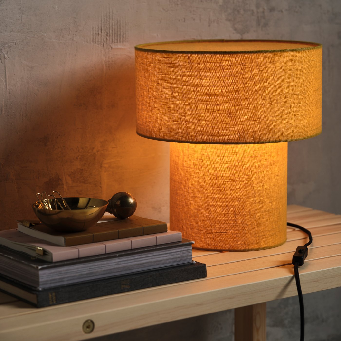 Närbild av en tänd STOCKHOLM bordslampa med gul lampskärm på en träbänk. Bredvid ligger staplade böcker och en liten skål i metall, vilket skapar en varm och mysig känsla.