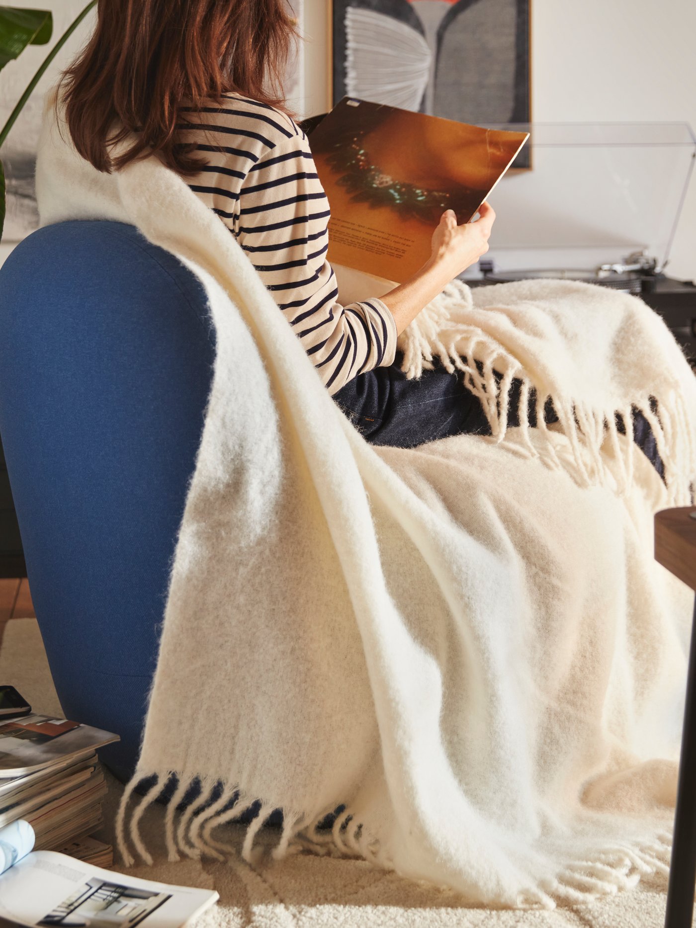 Een persoon die zit op een Gunnared donkerblauwe LILLESÄTER draaifauteuil met een zachte KLIPPOXEL plaid, naast een stapel tijdschriften.