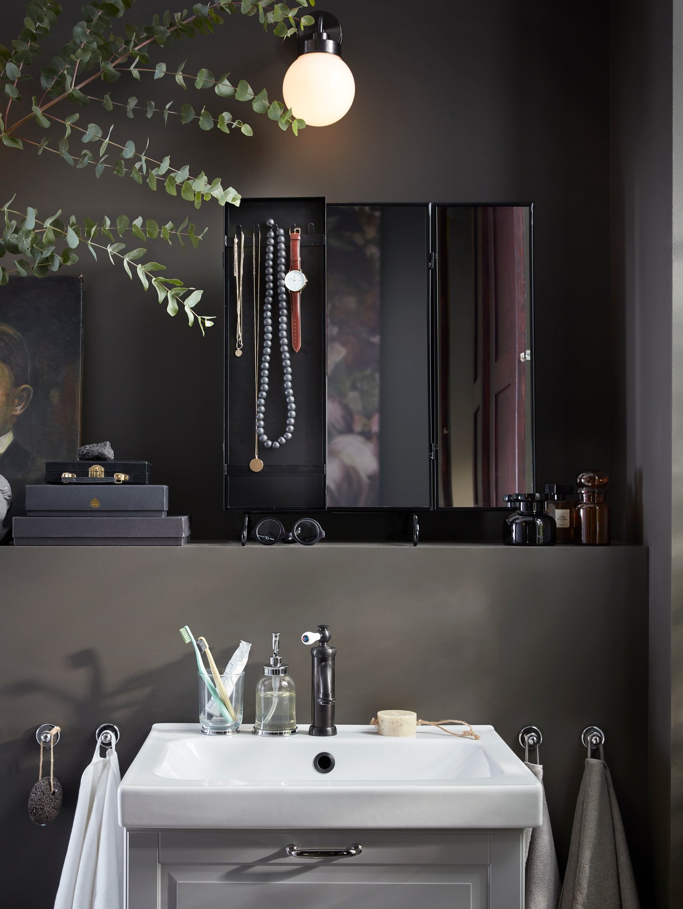 Bilik mandi berwarna kelabu gelap dengan cermin berwarna hitam pada para-para di atas sink cuci tangan, barang kemas dan aksesori digantung pada cermin.