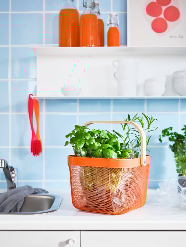 A kitchen top with herbs in an orange RISATORP basket. A white shelf on the kitchen wall holds cups, bowls and juice bottles.