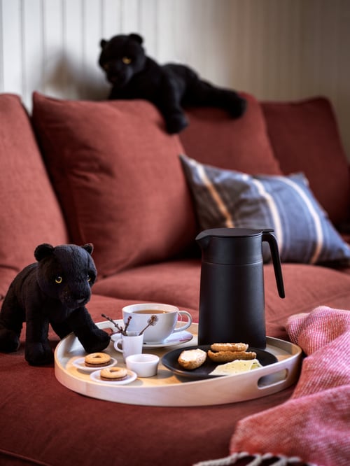 Close-up of a black UNDERLÄTTA vacuum flask on SKALA round tray with sweet treats; the tray is nestled on a red sofa.