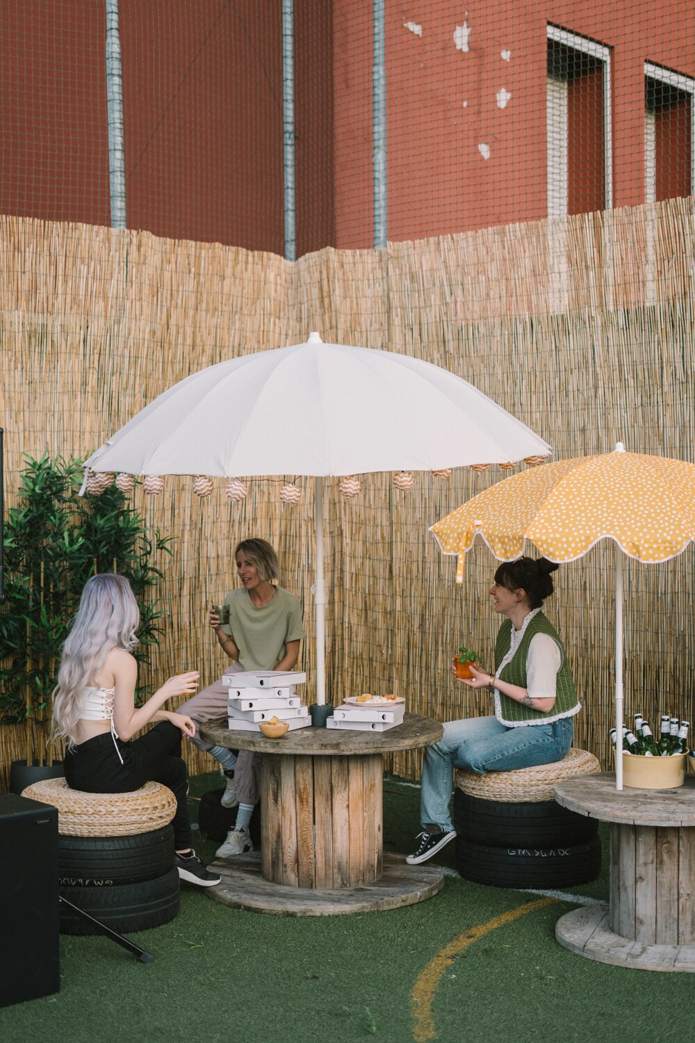 Tre donne che conversano all'ombra di ombrelloni SAMSÖ sedute su pouf e gomme di macchina. 