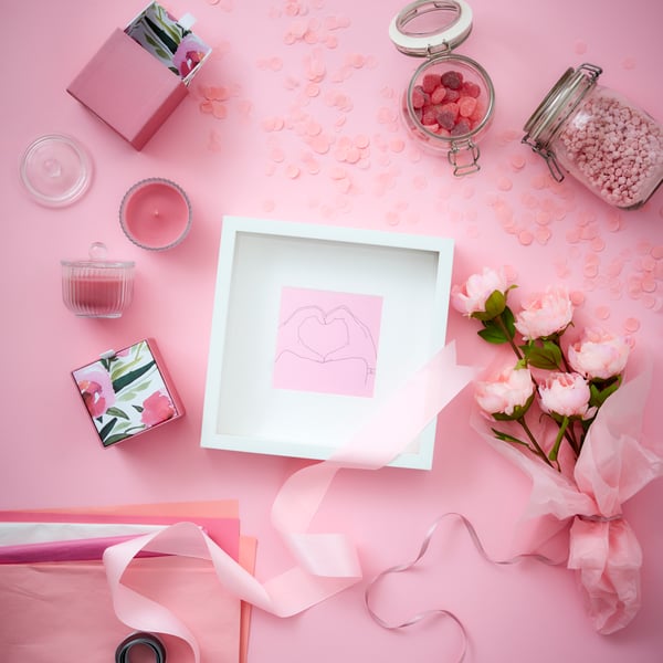 White square picture frame with a pink heart lying flat on a pink background with flowers, candles and candies nearby.