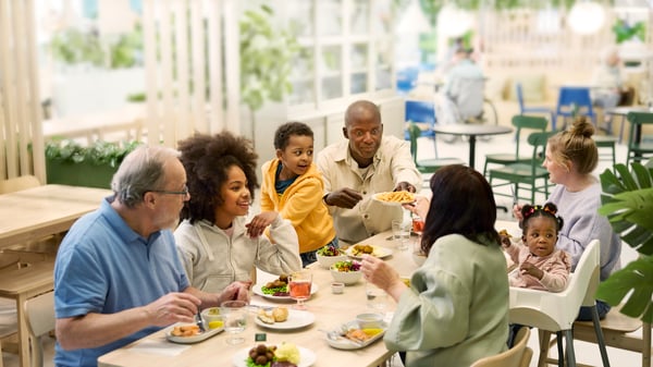 Eine Gruppe von Menschen unterschiedlichen Alters sitzt gemeinsam an einem Esstisch im IKEA Restaurant und genießt eine Mahlzeit.