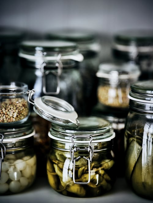 A shelf of KORKEN jars in different sizes filled with pickles, vegetables and spices. A jar at the front has the lid open.