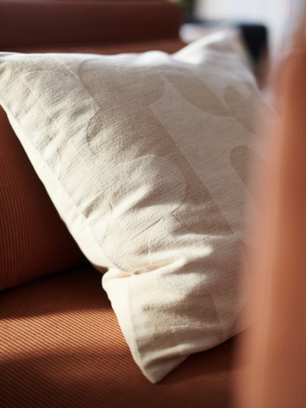 A beige RÖDASK cushion cover with a patterned surface on top of a rust-coloured sofa, in a sunny room.