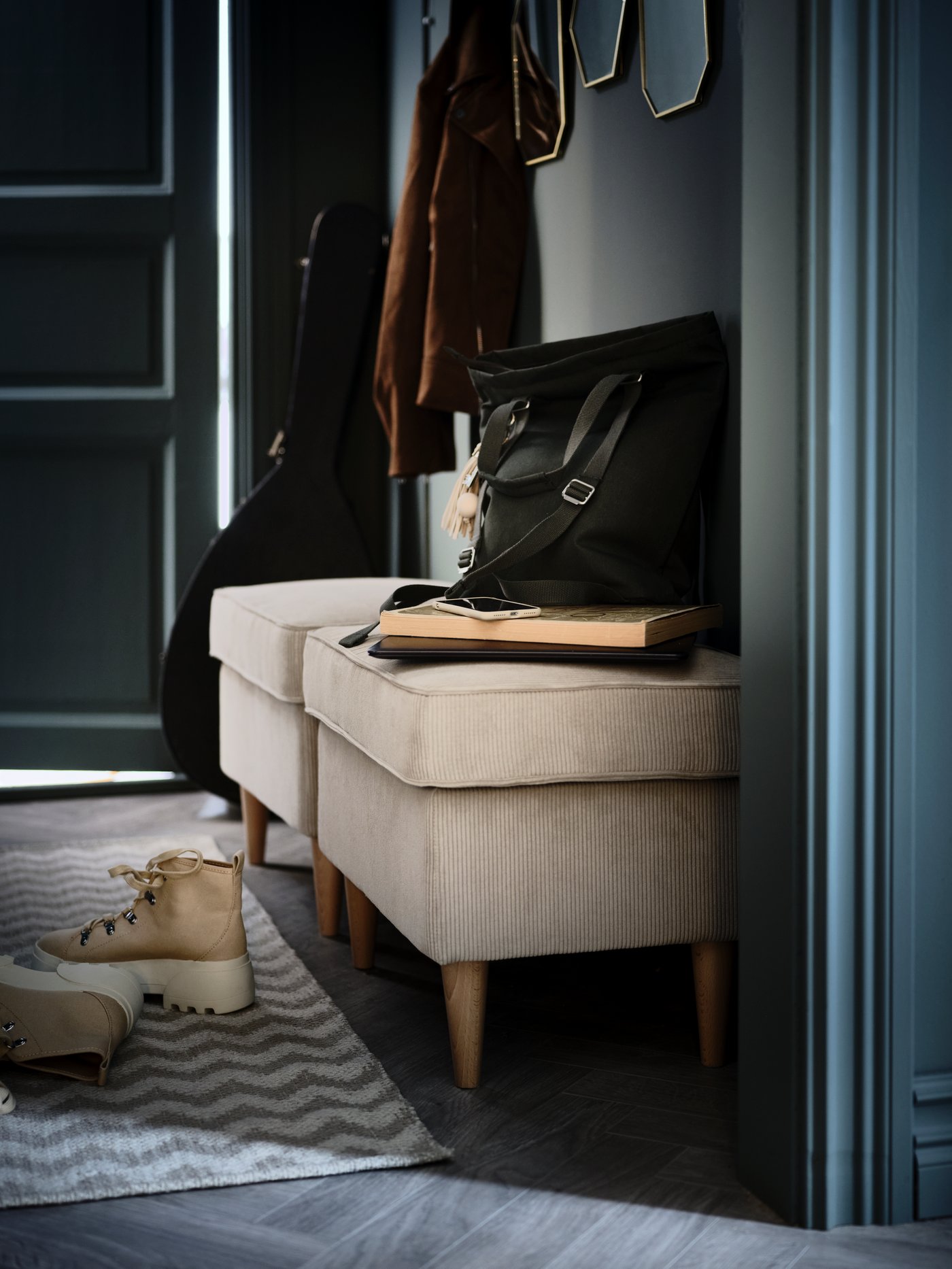 Dos poufs STRANDMON beige en la entrada. Debajo hay un par de botas sobre una alfombra BREDEVAD de color beige con estampado de zigzag.