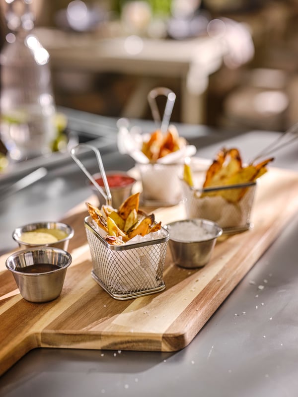 An acacia‑wood SMÅÄTA chopping board with stainless steel GRILLTIDER sauce cups and a stainless steel frying basket filled with fries.