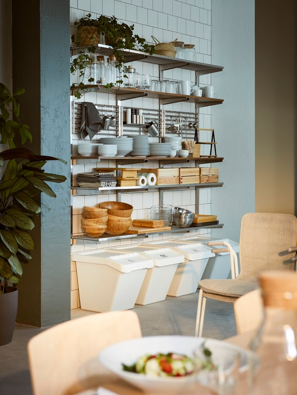 A row of SORTERA waste sorting bins placed by a white-tiled wall fitted with KUNGSFORS shelves laden with tableware.