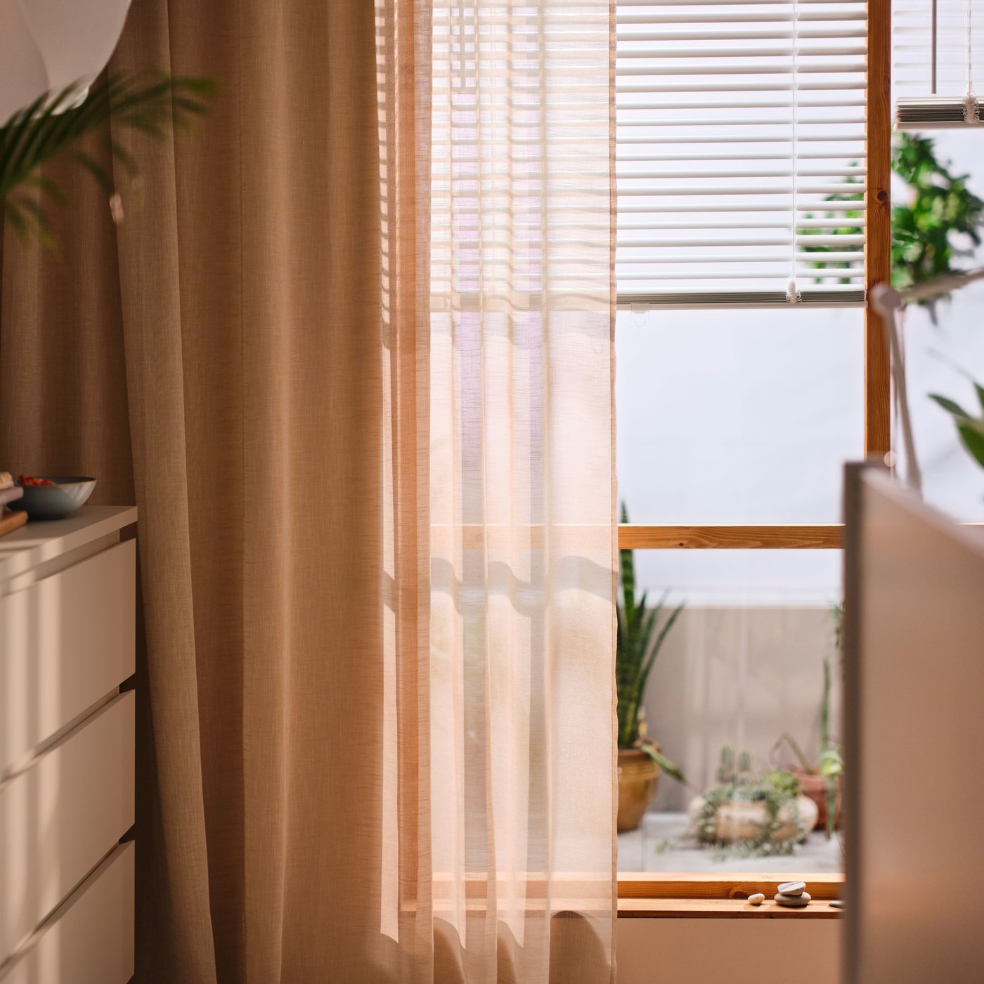 A white VECKLARFLY venetian blind half open on a window with a HÄLLEBRÄCKA sheer curtain in light beige in front of it.