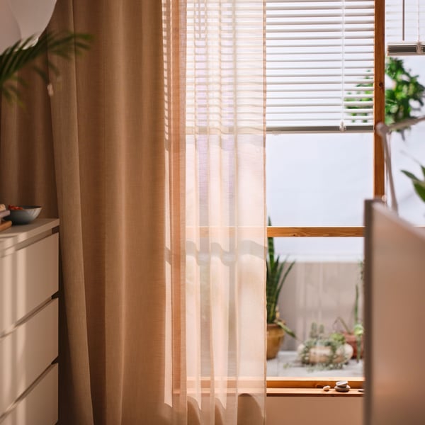 A white VECKLARFLY venetian blind half open on a window with a HÄLLEBRÄCKA sheer curtain in light beige in front of it.