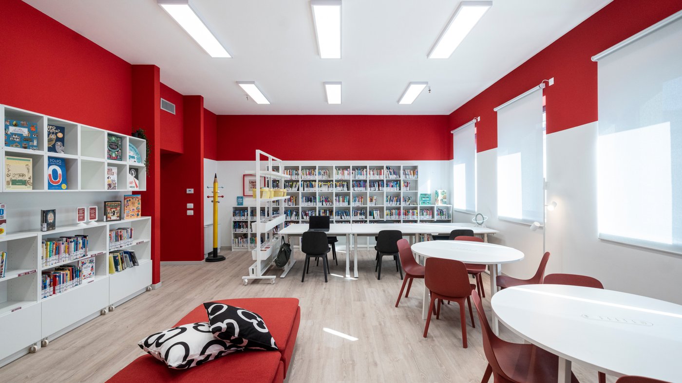 Vista di una stanza della biblioteca a cui lati ci sono un sistema componibile di mobili da parete EKET, libreria BILLY e una fila di tavoli e sedie rosse MIZZON. In mezzo due divanetti rossi. In fondo alla stanza è appoggiata alla parete una libreria BILLY