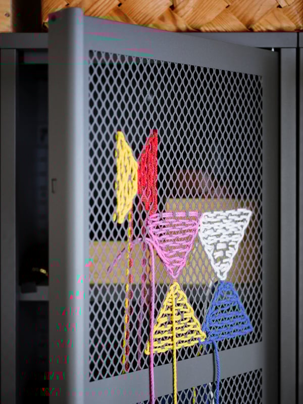 A section of a grey IVAR cabinet where colourful threads have been used to embroider decorations on the door's metal mesh.