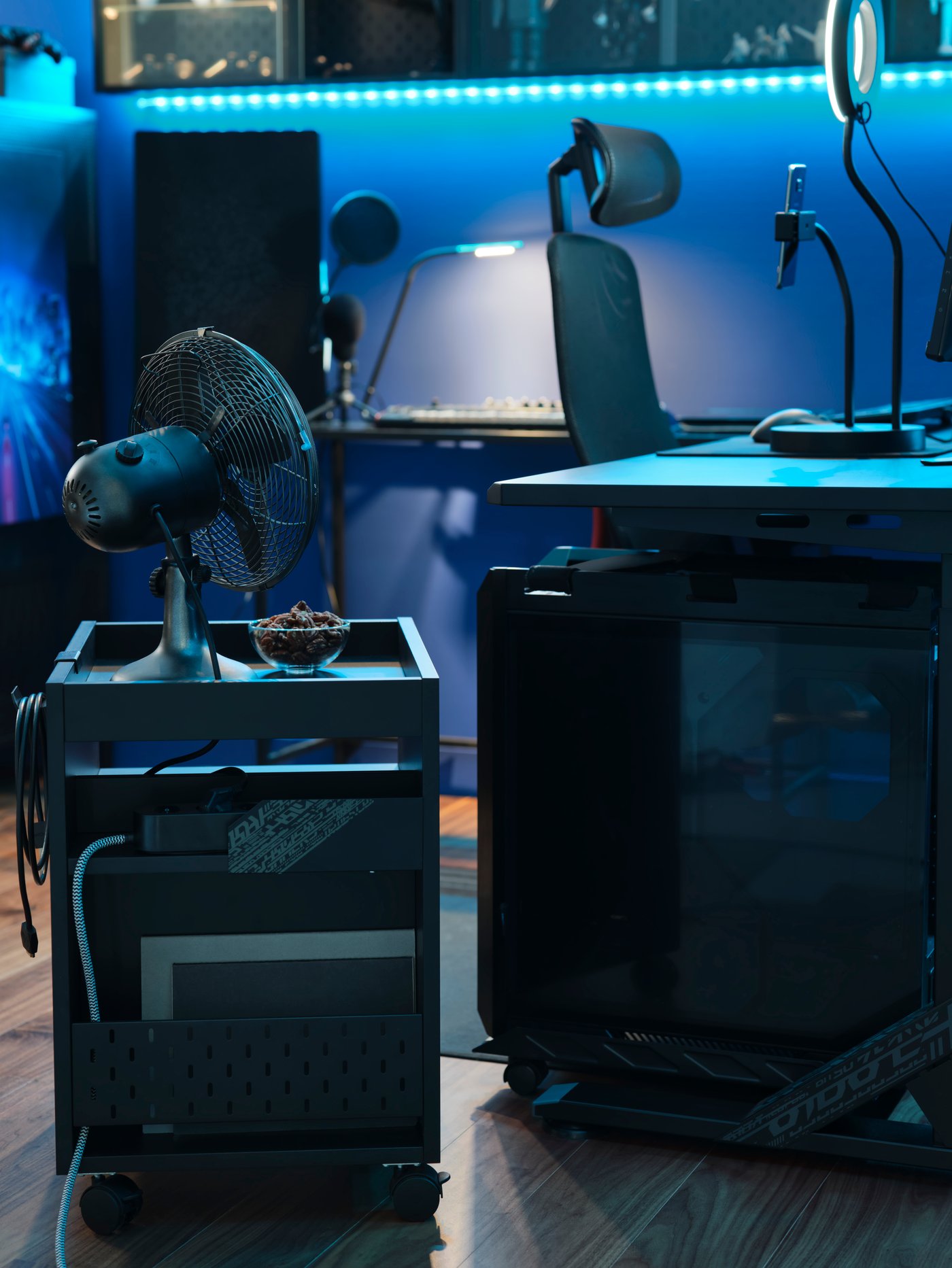 An UPPSPEL CPU stand and drawer unit by a desk in a blue-lit gaming room. There’s a fan and snacks on the drawer unit.