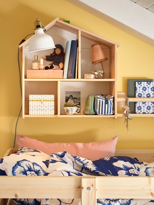 An einer Wand über einem Etagenbett ist ein FLISAT Puppenhaus/Wandregal aus Kiefer zu sehen, in dem sich verschiedene Spielsachen, Bücher, eine Leuchte, ein Stofftier und weitere Kinderartikel befinden.