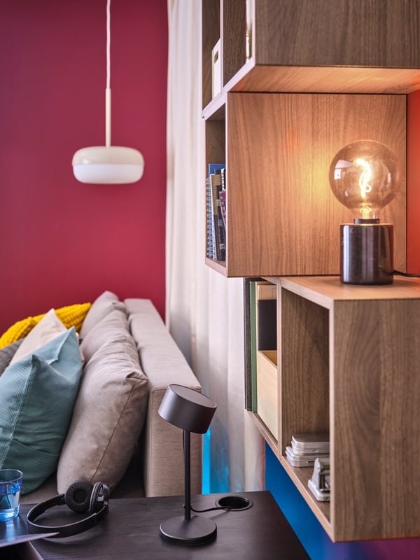 A black LÖVMÅNAD work lamp on a black desk, a white BLÅSVERK pendant lamp, a piece of a grey sofa with pillows.