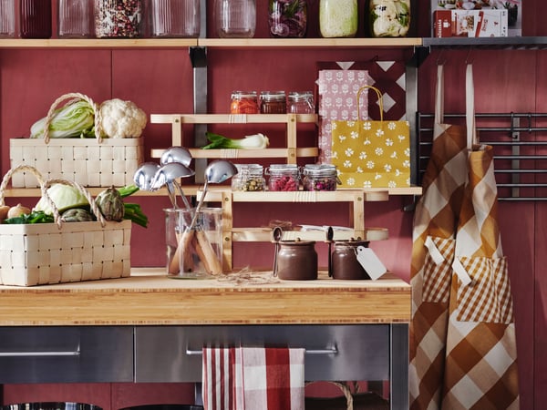 A kitchen with a RIMFORSA work bench made of stainless steel and bamboo, featuring various KÖSSEBÄR items such as jars, baskets, aprons, tea towels, and more.