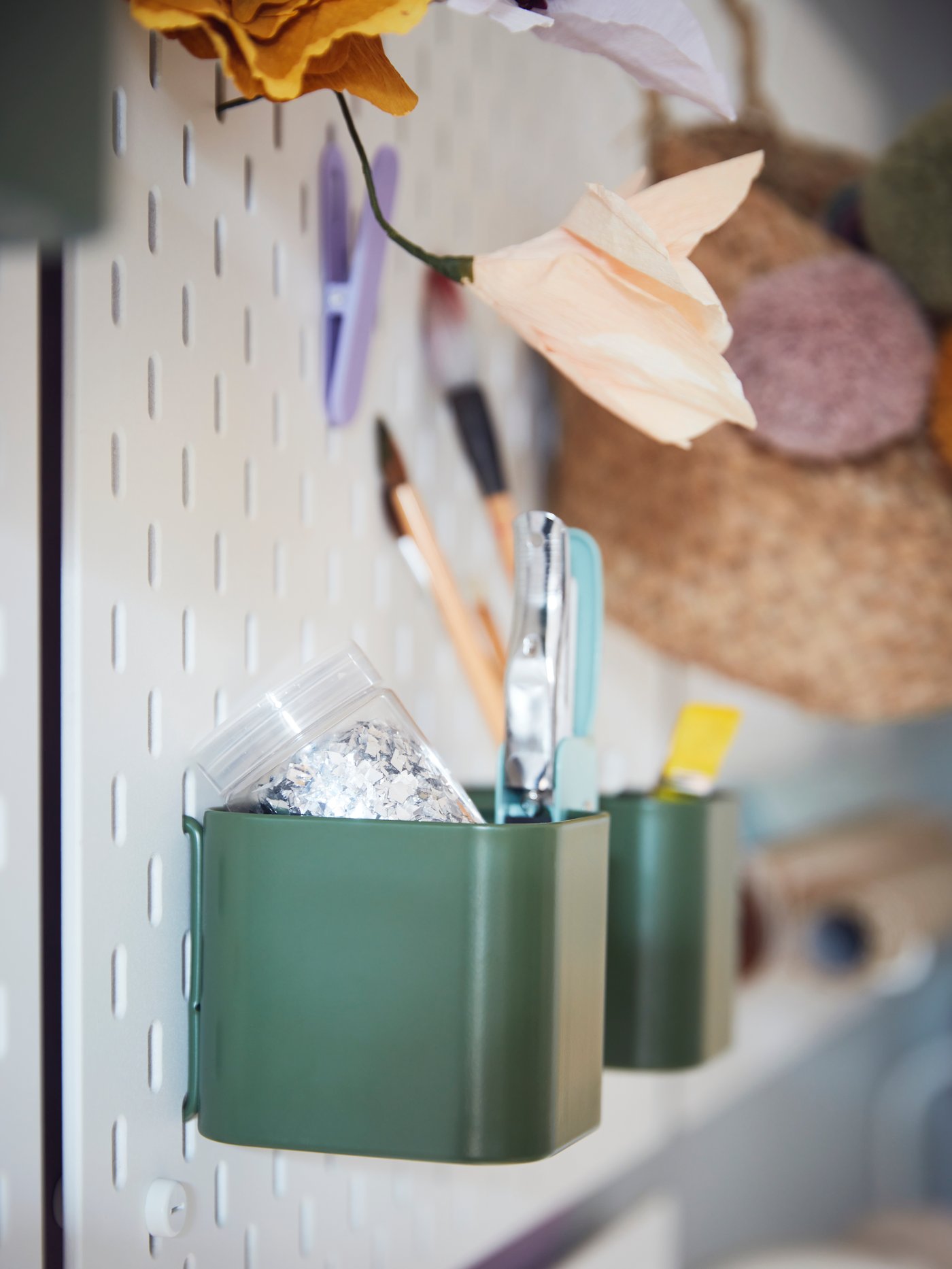 Parte di un pannello portaoggetti SKÅDIS bianco con ganci SKÅDIS lilla e contenitori SKÅDIS verde scuro per gli accessori da scrivania.