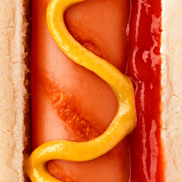 Primo piano di un hot dog IKEA nel panino, con una spirale di senape gialla e una striscia di ketchup rosso: pochi ingredienti, tanto gusto, pronto da addentare.