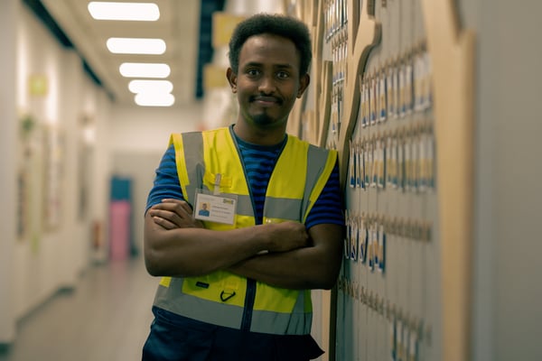 Abdi, wearing a high-visibility vest, stands with arms crossed in a corridor, smiling.