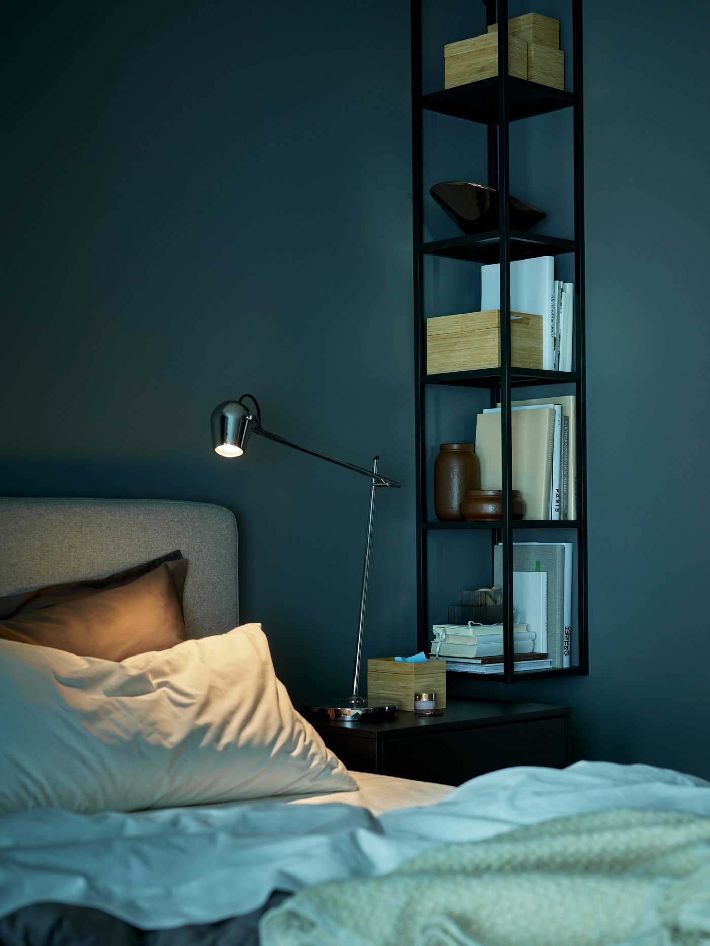 Hörnet av en säng med ett ENHET hyllsystem på väggen och en MODERMOLN arbetslampa på sängbordet. Rummet är mörkt, som på natten.