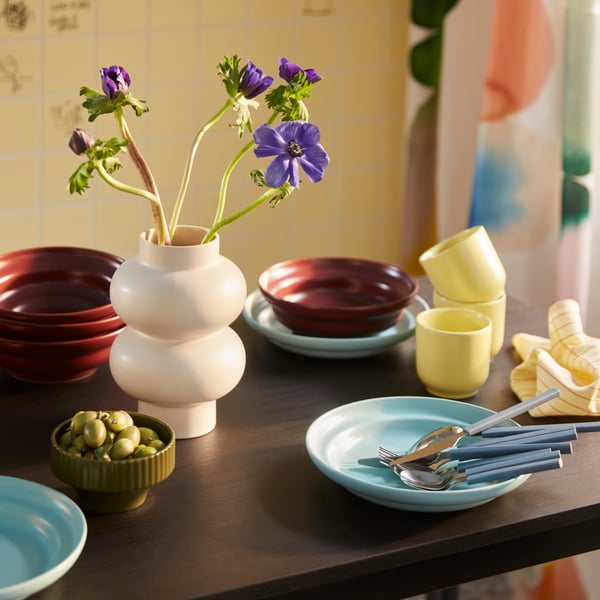 A dining table set with SPJUTLILJA serveware including a dark purple bowl, a yellow cup and a light blue plate.