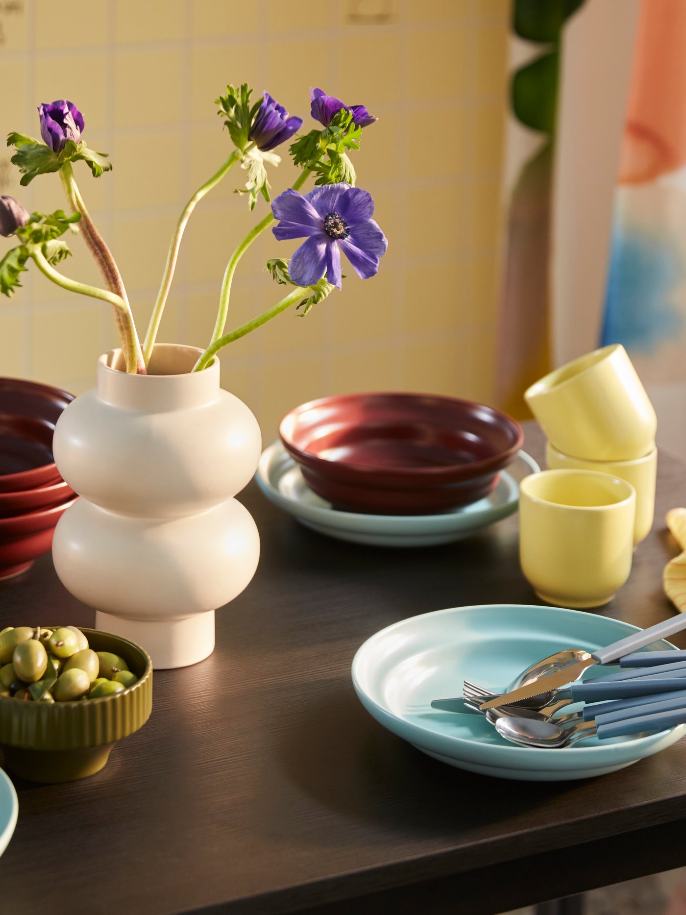 A dining table set with a SPJUTLILJA three-piece serving set, including a dark purple bowl, a yellow cup and a light blue plate.