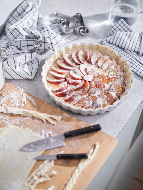 A worktop with FÖRDUBBLA knives and dough slabs on a LÄMPLIG chopping board, next to an unbaked peach pie in a VARDAGEN dish.