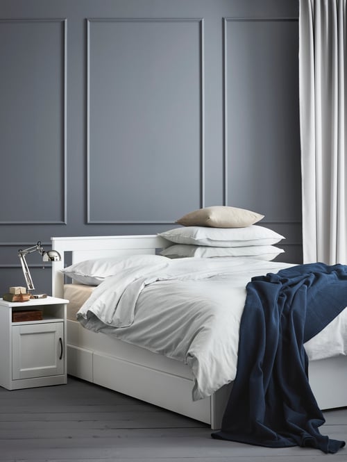 Bedroom with a bed frame with bedspread and stacked pillows, with a bedside table with work lamp to the side.